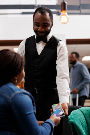 Cashless hotel. African American woman guest making contactless payment using credit card while resting in lobby. Smiling waiter holding pos terminal accepting payment over nfc technologyの写真素材
