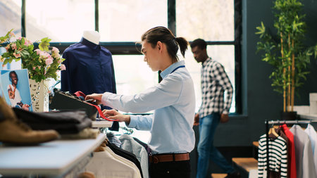 Asian worker arranging stylish tie, putting accessories on shelf in modern boutique. Retail employee preparing store for opening, working with fashionable clothes in shopping mall. Fashion conceptの写真素材