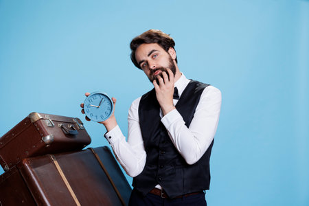 Male hotel porter posing with clock on camera, expressing punctuality and hating to be late in tourism industry. Doorman employee looking at time on wall watch, trolley bags.の写真素材