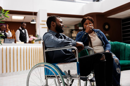 Young African American man on wheelchair sitting in hotel lobby with wife, tired unhappy woman travelling with disabled husband. Inclusive hospitality, handicap people and travelの写真素材
