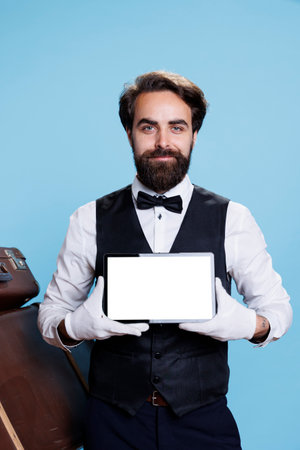 Classy hotel porter with white screen on tablet, posing on camera next to pile of baggage. Young man bellhop presenting blank gadget display, showcases professionalism of his occupation.の写真素材