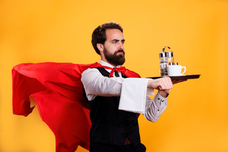 Elegant waiter with red cape carrying coffee on tray, having towel hanging over hand and posing as a superhero in studio. Young adult restaurant butler serving food and drinks.の写真素材