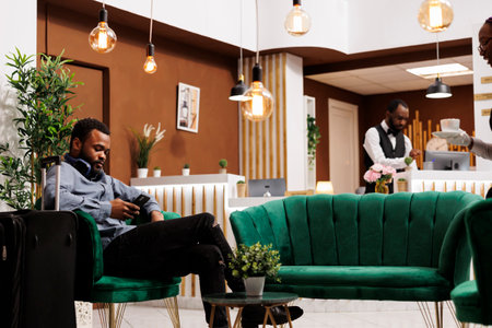 Black guy traveler sitting in hotel lobby with smartphone making mobile check-in. Young African American man tourist with luggage using phone texting sms message, waiting for roomの写真素材