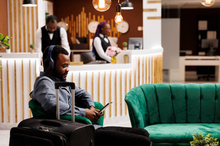 Smiling young African American man traveler waiting for room in hotel. Black guy in headphones using digital tablet while sitting in lobby with luggage, watching movie, wait for check-in processの写真素材