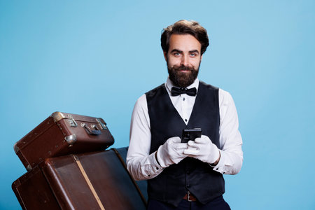 Hotel porter scrolls on smartphone, using social media and texting messages while he poses in studio with trolley bags. Doorman bellhop in suit browsing online page on phone app.の写真素材