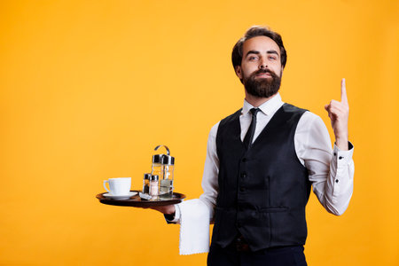 Waiter with serving tray points upwards on camera, showing something up above head while he wears professional restaurant clothes. Young man butler suggesting direction in studio.の写真素材