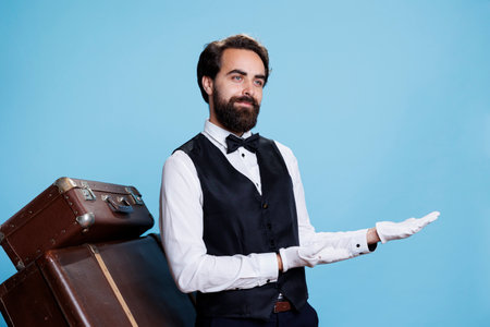 Hotel porter shows left side on camera, wearing professional suit and gloves while he does an advertisement, suggesting direction sideways. Elegant doorkeeper presents ad in studio.の写真素材
