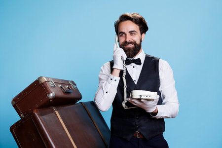Visitors calls hotel telephone and discuss with refined bellboy about bookings, studio background. Skilled hotel receptionist uses corded phone to talk about commodities and concierge services.の写真素材