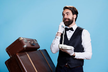 Stylish bellboy answers landline phone in studio, talking to hotel guests about accommodation. Skilled confident doorman using telephone with cord to take calls, vintage luxurious job.の写真素材