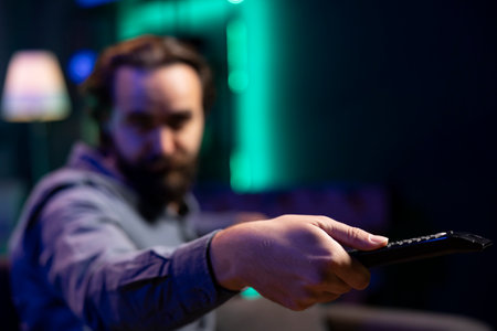 Focus on remote control used by man in blurry background switching channels on television screen. Close up of device used by person to scroll through streaming content, picking movieの写真素材