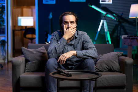 Viewer gasping while watching horror television series on streaming service, enjoying time off from work. Astonished person covering mouth in disbelief, shocked by film scene broadcasted on TVの写真素材