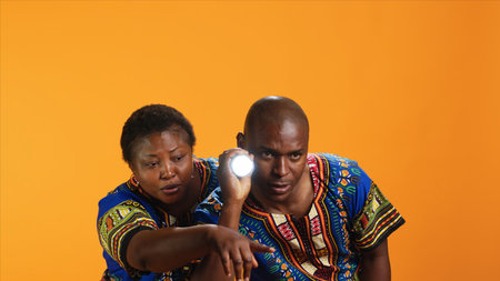 Confident man using flashlight to see what caused noise, trying to calm his wife who is hiding behind him. Terrified woman being anxious about weird sound, husband using lamp light and searching.の写真素材