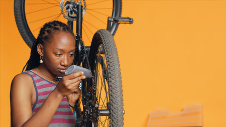 African american woman using specialized glue to repair broken bicycle chain, studio background. Skilled engineer applying adhesive on bike components during maintenance process, camera Aの写真素材