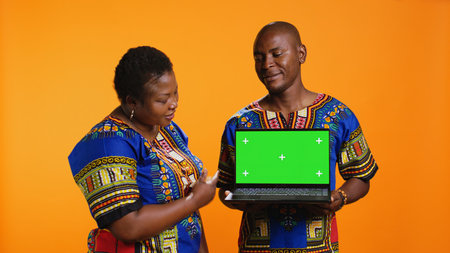 African american partners holding laptop with greenscreen on camera, presenting isolated display with chromakey and standing over orange background. Man and woman showing blank mockup.の写真素材