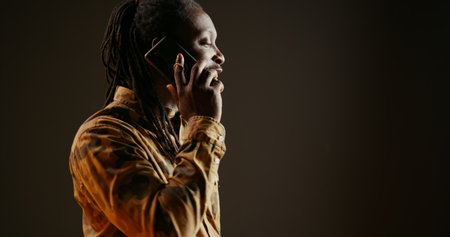 Cool guy with dreads answering phone call in studio, talking to friends or family on camera. African american trendy adult chatting remotely with people on telephone line. Handheld shot.の写真素材