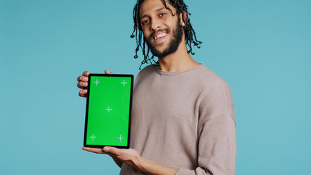 Joyful man holding green screen tablet, showing positive emotion. Upbeat person presenting chroma key device, doing recommendation, isolated over studio background, camera B close upの写真素材