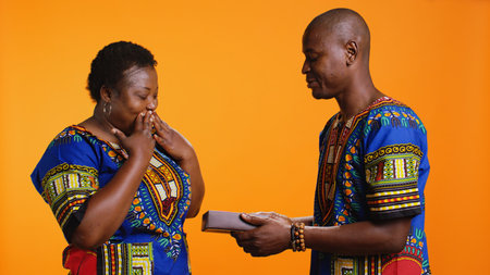 Romantic man offering present with ribbon and bow to his wife, giving giftbox to celebrate her birthday or their anniversary. African american woman receiving sweet wrapped gift from her partner.の写真素材