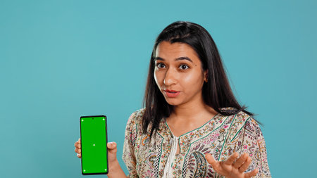 Woman presenting green screen mobile phone, isolated over studio background. Cheerful indian person holding copy space chroma key smartphone used for advertising brands, camera Bの写真素材