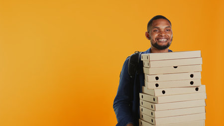 Male pizzeria courier carrying huge pile of pizza boxes in studio, preparing to deliver meal order to customers. Young deliveryman holding a big stack of fast food takeaway, shipping. Camera B.の写真素材