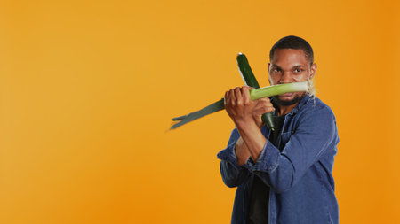 Pleased young adult pretending to start a fight with a cucumber and a leek, acting silly having fun in studio. Vegan guy playing around with natural eco friendly green vegetables. Camera B.の写真素材
