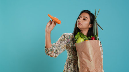 Woman with paper bag in hands filled with vegetables checking carrot for impurities. Conscious living customer with purchased groceries to be used as cooking ingredients, studio background, camera Aの写真素材