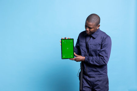 Trained armed bodyguard holds a tablet with greenscreen layout on camera, presenting isolated copyspace against blue background. Security officer posing in uniform with device in studio.の写真素材