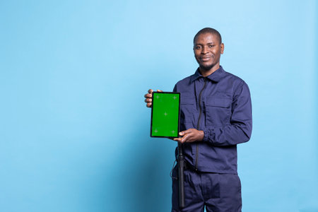 Security officer holding a device with greenscreen display in studio, posing with safeguarding uniform on camera. Guard presenting a tablet with greenscreen layout, uniform equipped with baton.の写真素材