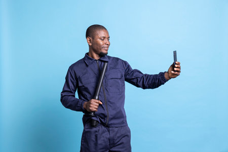 Smiling security agent taking photos with his baton on camera, using a smartphone to take pictures against blue background. Young guard makes memories in his patrol uniform in studio.の写真素材