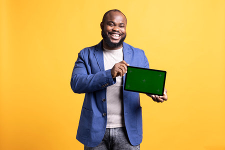 Happy african american laughing while man holding green screen tablet, doing recommendation. Overjoyed BIPOC person presenting chroma key device, isolated over studio backgroundの写真素材