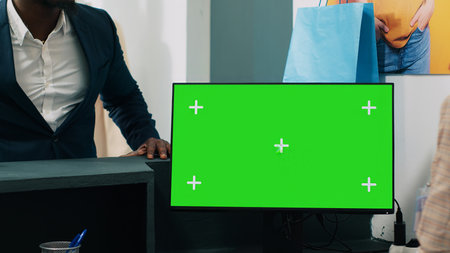 African american worker uses computer with greenscreen display on at checkout counter, putting clothes in a bag for customer. Employee looks at pc showing copyspace mockup screen. Camera A.の写真素材