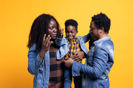 African american family of three smiling against yellow background, posing with affection and gratitude. Cheerful young parents holding their cute infant son, enjoying time together.の写真素材