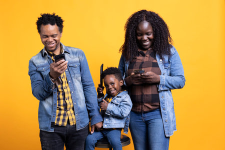 African american parents checking their smartphones next to the kid on camera, adorable young boy playing with a remote control. Happy family having fun posing with electronics in studio.の写真素材