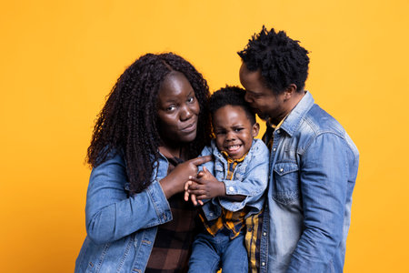 African american family and their little child in studio, holding each other against yellow background. Smiling proud young couple posing with their adorable toddler, enjoying leisure time.の写真素材