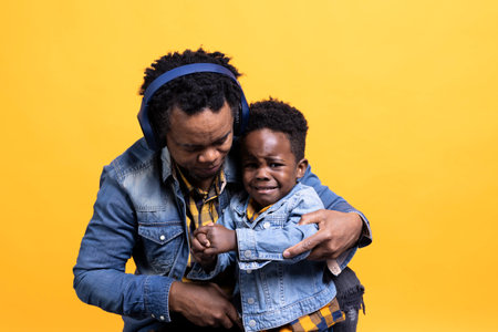 Sad little baby boy starts crying and his dad is holding him, trying to calm him down while he wears headphones. Displeased unhappy toddler cries against yellow background in studio.の写真素材
