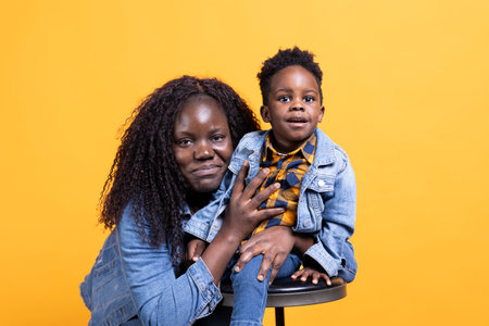 Proud mom and young toddler smiling and posing in front of the camera, adorable small child is sitting on a chair next to his mommy. African american little family shows affection in studio.の写真素材
