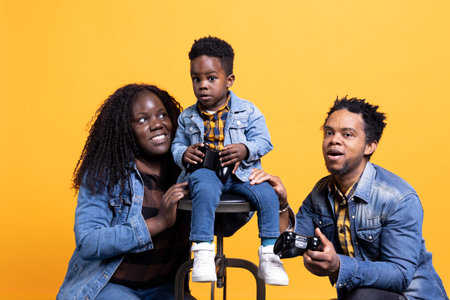Cheerful satisfied father playing video games with his toddler using a joystick, young parents having fun with their small boy. African american loving father plays with kid online.の写真素材