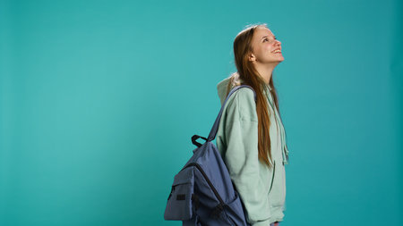 Joyous girl holding rucksack looking around, inviting others to join her, doing hand gesturing. Jolly teenager doing inspecting gesture, discovering new place, isolated over studio backdrop, camera Bの写真素材