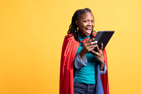 Portrait of ecstatic african american girl portraying superhero excitedly using tablet, studio background. Delighted woman posing as hero in costume, using digital device, having funの写真素材