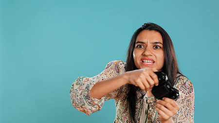 Upset woman showing thumbs down sign gesturing holding controller, after being defeated in videogames. Displeased indian person doing rejection hand gesture after losing, studio background, camera Bの写真素材