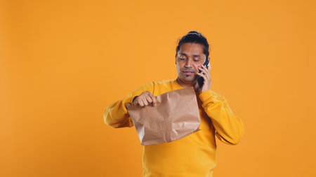 Delivery man arguing with customer on phone after arriving at destination and waiting for him, studio background. Indian person doing deliveries, scolding late client on telephone call, camera Bの写真素材