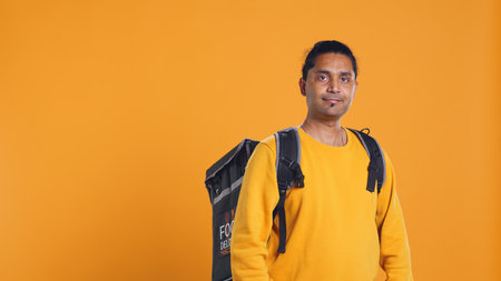 Portrait of happy smiling indian bike rider holding thermal backpack, isolated over studio background. Friendly courier prepared to deliver takeaway food to customer, camera Bの写真素材