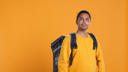 Portrait of confident indian man holding thermal backpack, crossing arms, studio background. Radiant nonchalant delivery person prepared to deliver takeaway food to customers, camera Bの写真素材