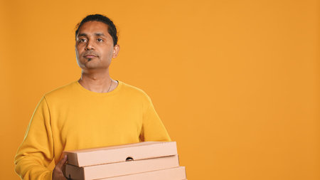 Portrait of courier isolated over studio background carrying stack of pizza boxes, close up shot. Indian employee delivering food waiting for client to answer door and pick up meal order, camera Aの写真素材