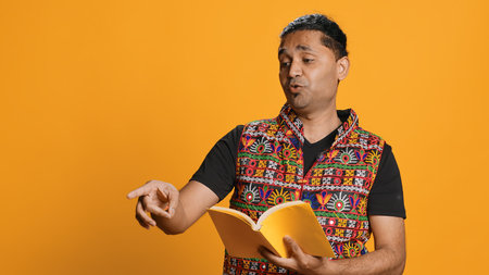 Joyous indian teacher reading book for pupils during class, isolated over studio background. Cheerful man holding novel, telling story to kindergarten kids, teaching them, camera Aの写真素材