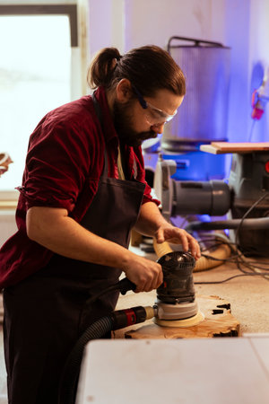 Carpenter wearing safety glasses, using orbital sander with finer sandpaper grits to achieve refined finish. Manufacturer using angle grinder on wood block, achieving professional quality resultsの写真素材