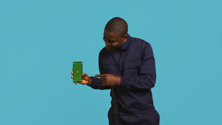 Portrait of joyous security guard at work showcasing green screen phone while overseeing premises. Jolly private law enforcement worker presenting mockup smartphone, studio backdrop, camera Bの写真素材