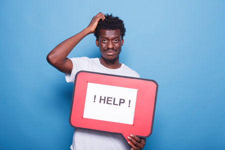 In studio, African American person is clutching a red speech bubble. Black man standing over isolated blue background while holding a speech board, asking for assistance.の写真素材