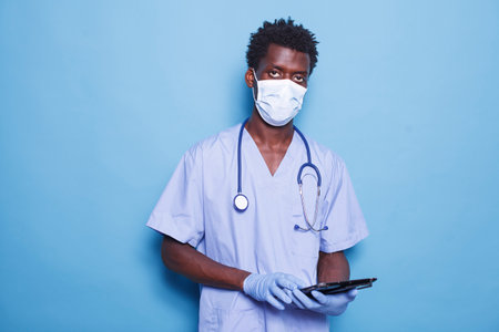 African American doctor in blue scrubs and face mask stands against a blue background while holding a tablet. Black man with stethoscope and gloves, grasping a digital device.の写真素材