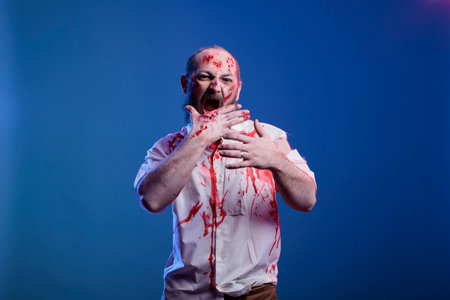Actor dressed as zombie acting for horror movie scene, pretending to eat human brain. Man costumed as undead monster wearing fake blood makeup consuming flesh, studio backgroundの写真素材