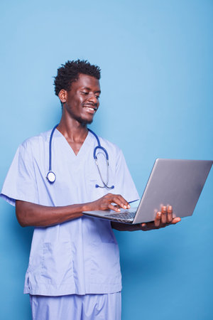 Smiling African American male doctor in scrubs standing and holding digital laptop. Black medical professional with stethoscope ready to provide healthcare by using wireless computer.の写真素材